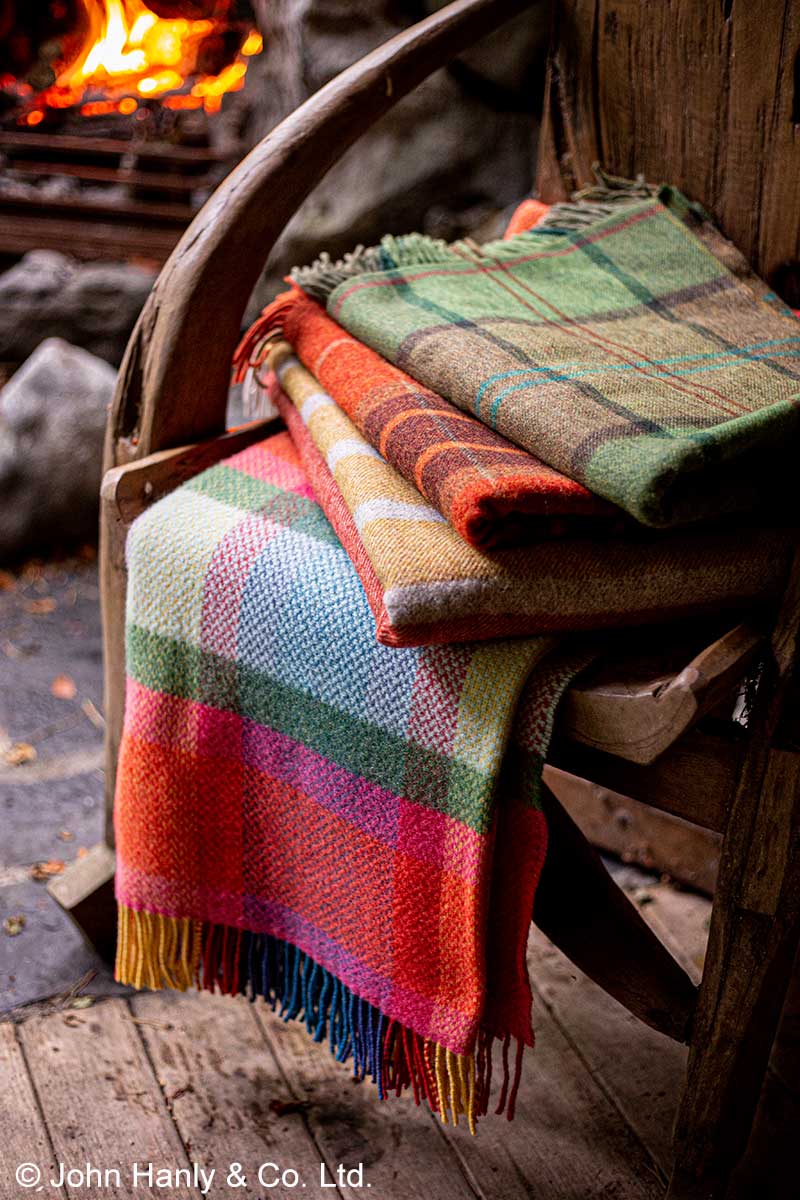 Vier verschiedene Plaids aus Wolle und mit Karomuster auf einem Holzstuhl, im Hintergrund brennt ein Kaminfeuer.